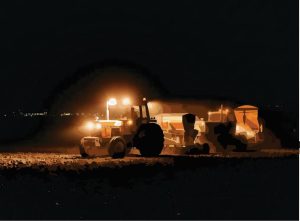 A farmer works on the field late at night with the help of some lights from the big rig